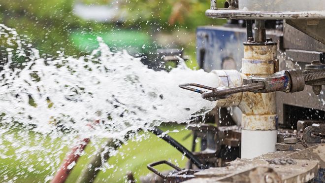 Water spraying from an industrial pipe or nozzle outdoors, with splashing droplets and machinery in the background.