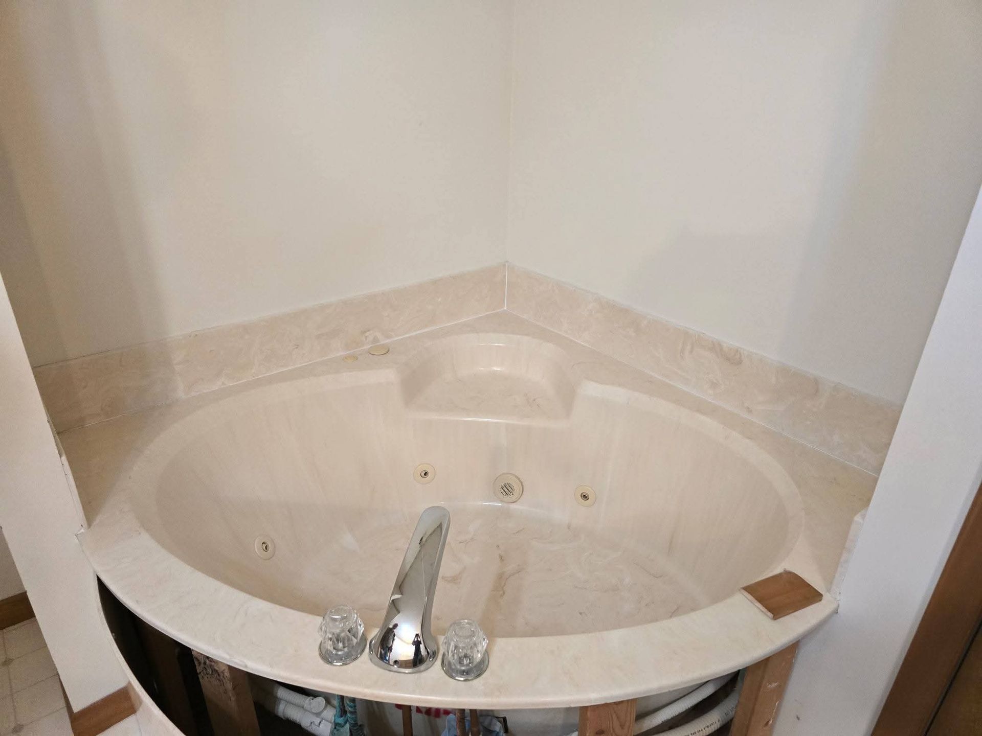 Cream-colored, corner jacuzzi tub with marble trim and chrome faucet, partially installed in a bathroom.