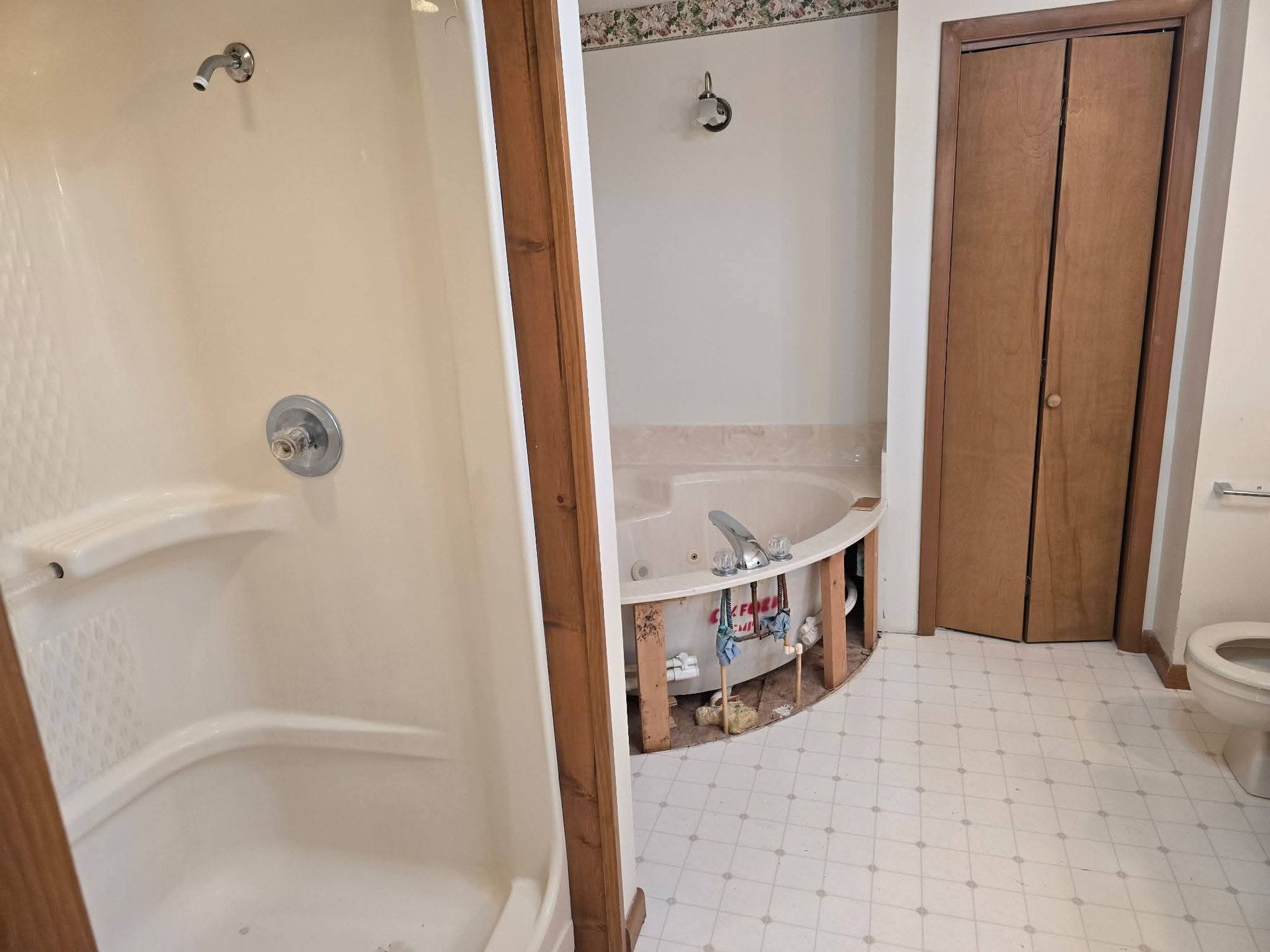 Bathroom with a shower, a partially dismantled tub, and a closed door. Light-colored walls and flooring.