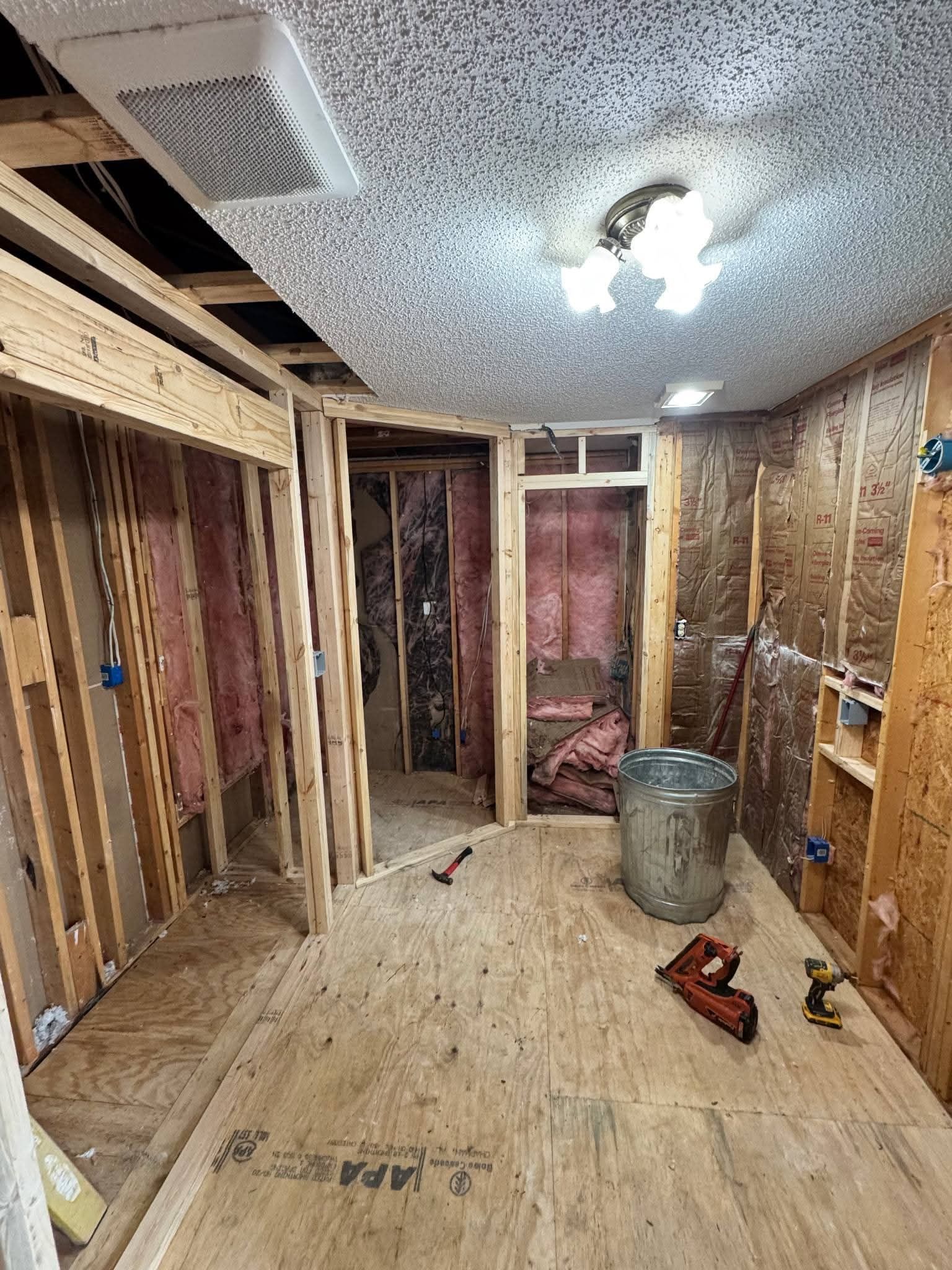 Bathroom under renovation, studs exposed, pink insulation, gray ceiling, tools on floor.
