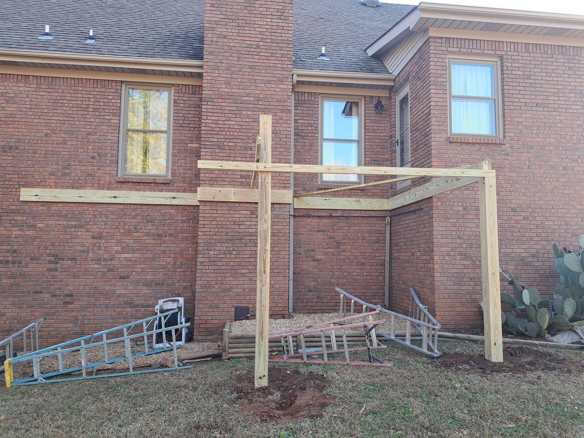 Wooden deck frame under construction against a brick house. Ladders and materials lie on the ground.