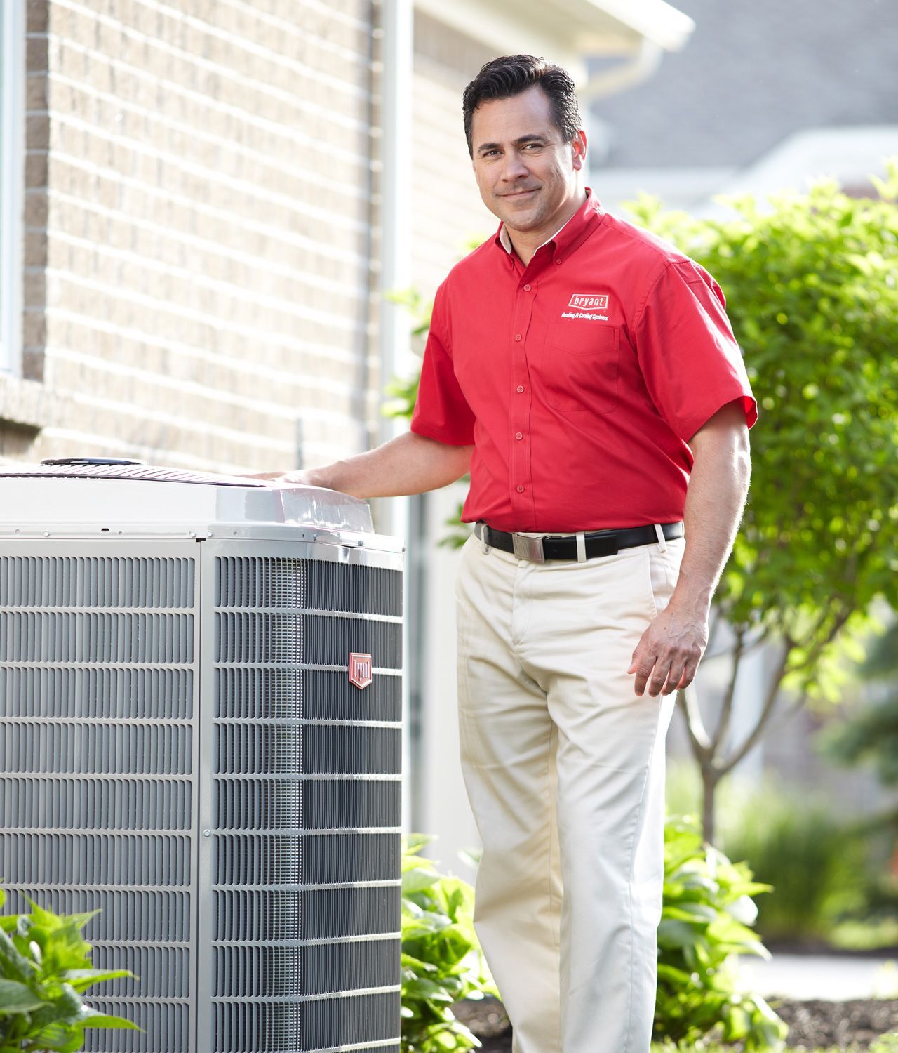 HVAC technician in a red shirt and khakis beside an air conditioning unit outside a house.