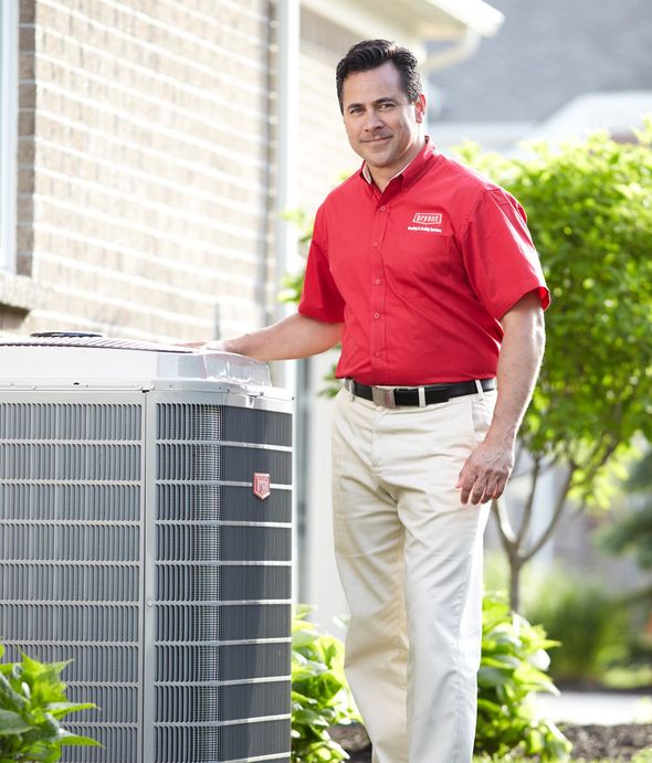 HVAC technician in a red shirt and khakis beside an air conditioning unit outside a house.