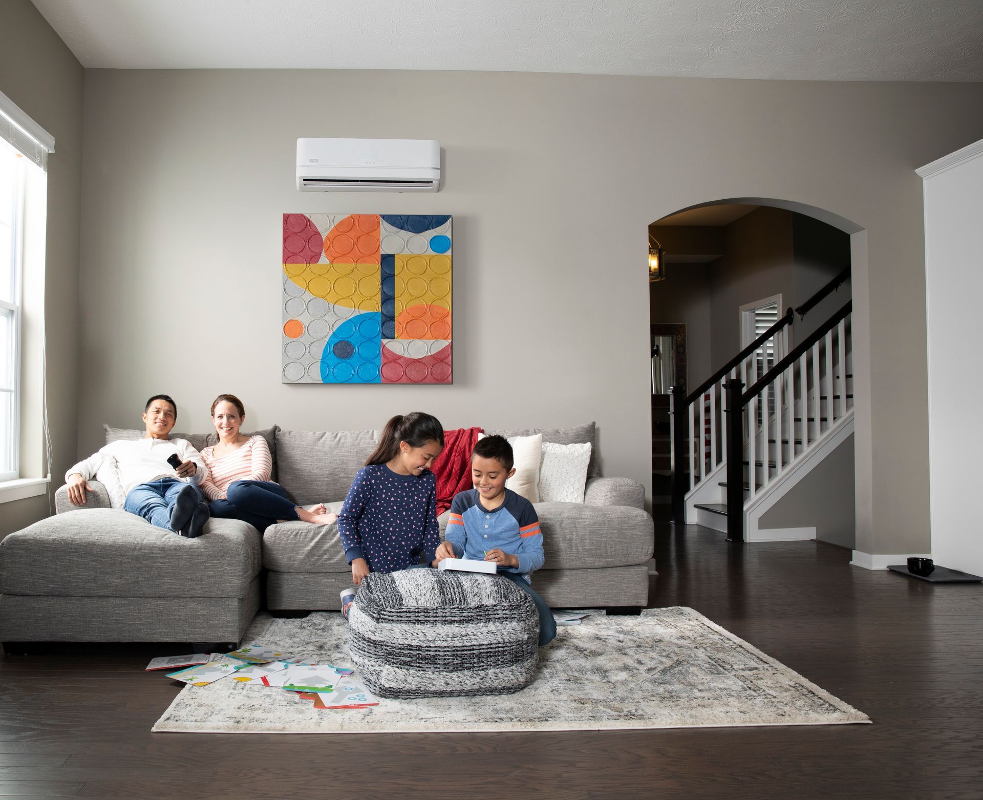 Family relaxing in living room; children drawing on ottoman. Air conditioner mounted on wall.
