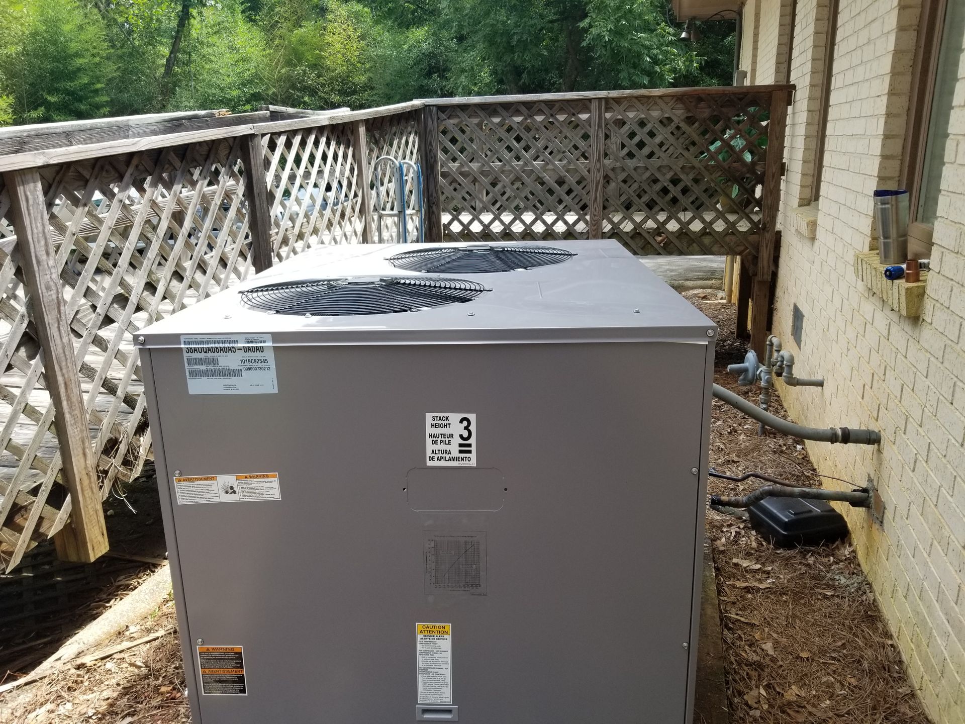 Gray air conditioning unit next to a light-colored brick building, with a wooden fence in the background.
