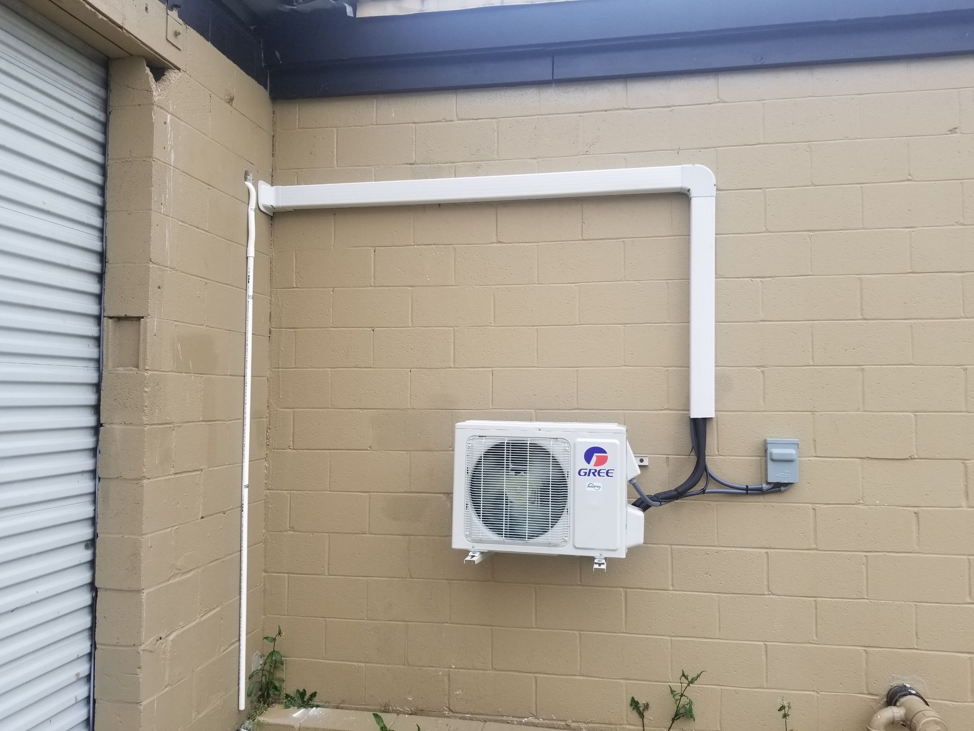 Wall-mounted air conditioning unit on a tan brick building with white conduit piping.