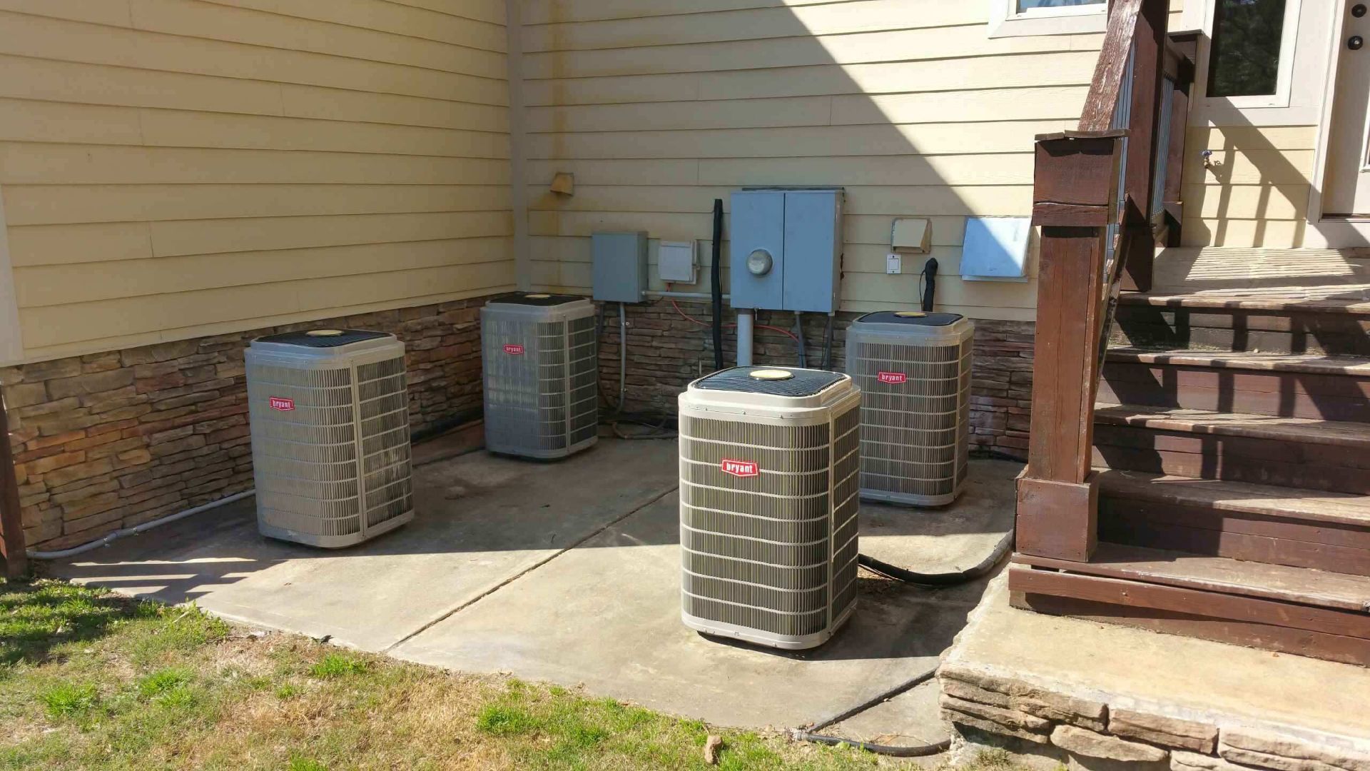 Several air conditioning units installed on a concrete slab against a building's wall with electrical boxes.