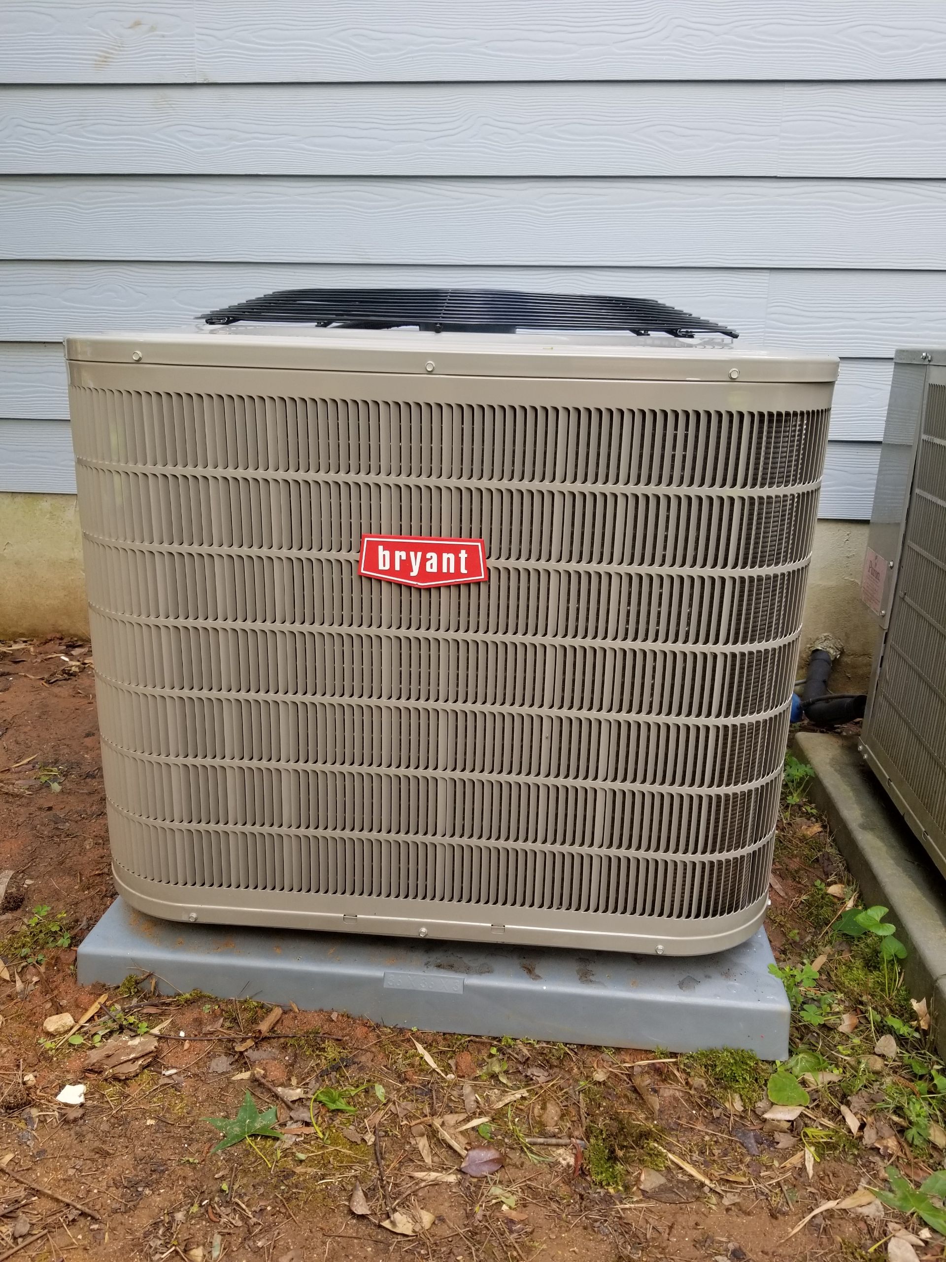 Outdoor Bryant air conditioning unit, tan and brown, on a gray pad, against a gray siding wall.