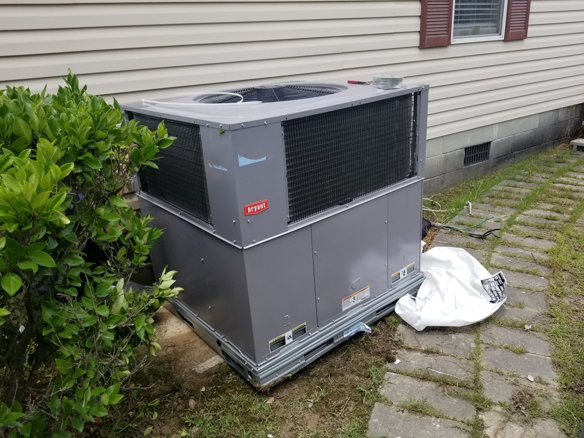 Gray HVAC unit next to a green bush and a tan house with a small window.