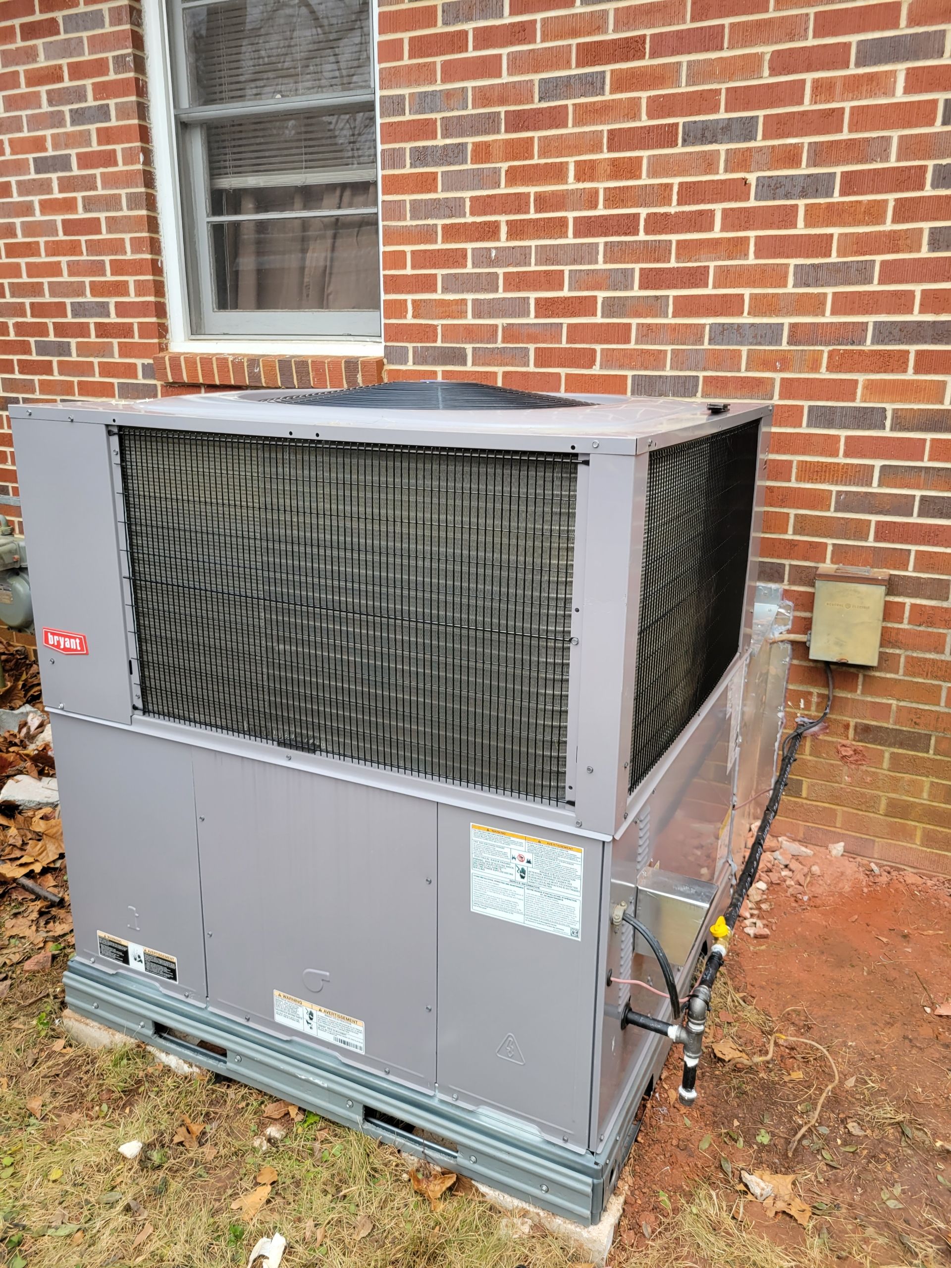 Gray outdoor HVAC unit against a brick building, near a window.