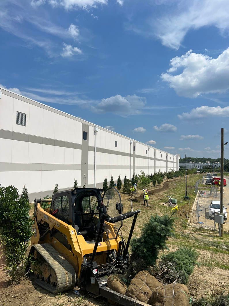 Yellow excavator at a construction site beside a long white industrial building under a blue sky