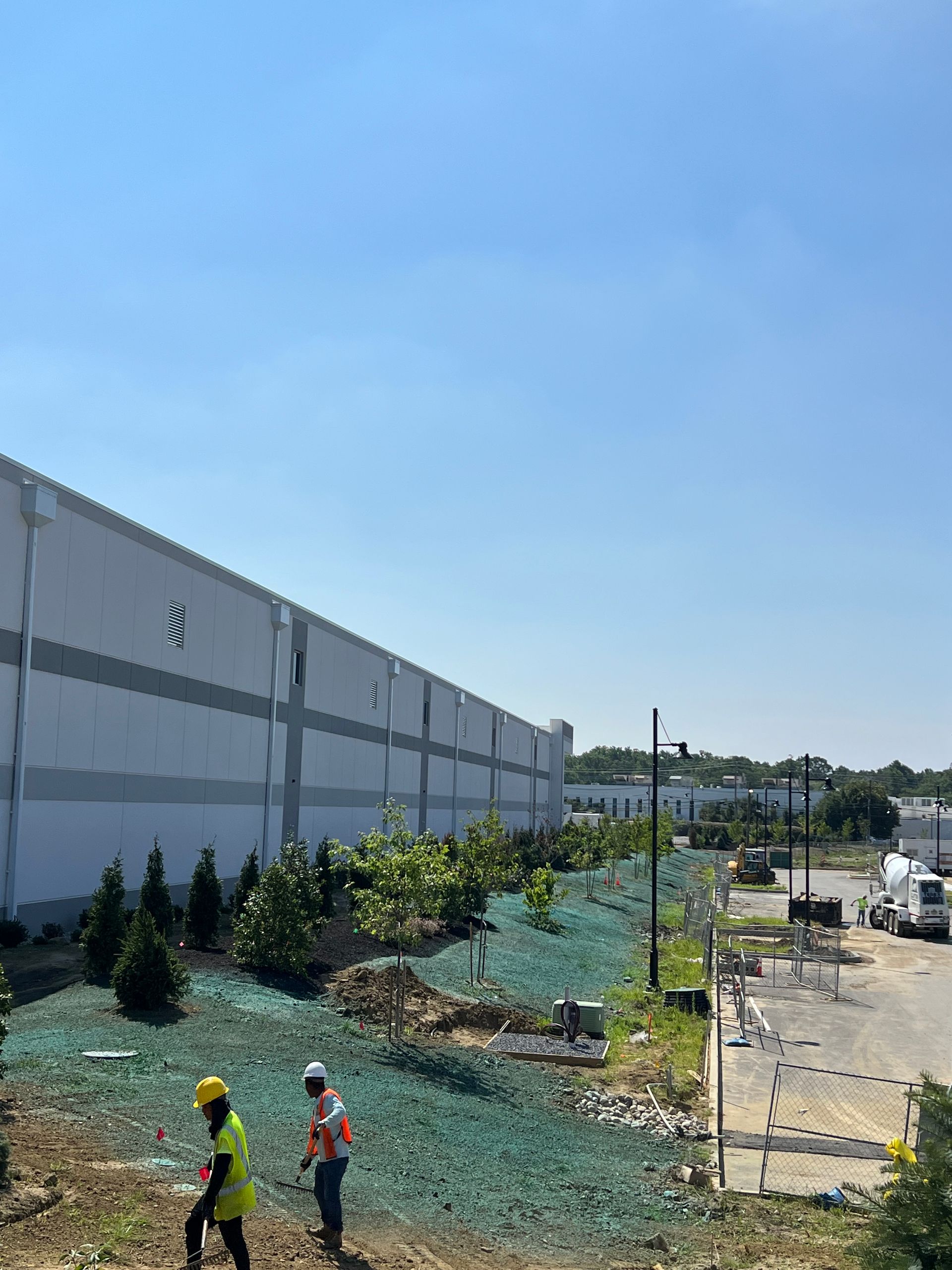 Construction workers near a long gray wall, landscaping a rocky roadside under a clear blue sky.