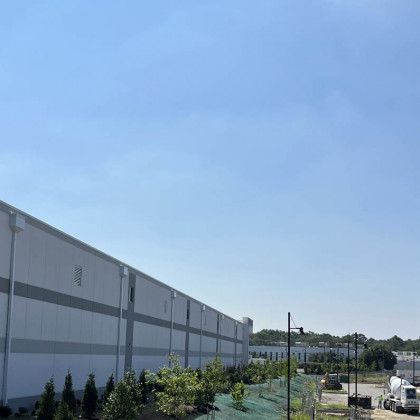 Industrial building with a long gray wall, landscaped shrubs, and parked vehicles under a clear blue sky