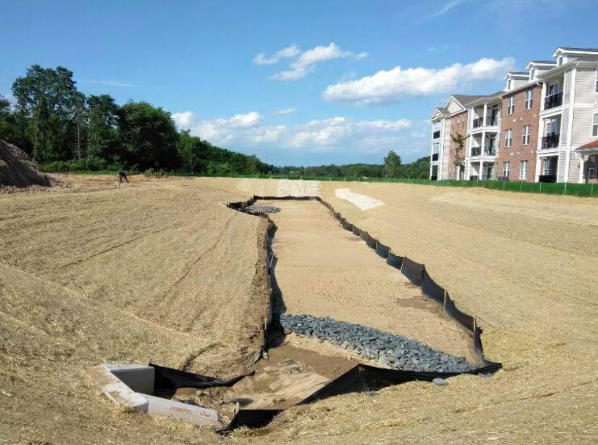 Large ground crack on a dirt field beside apartment buildings and trees under a blue sky