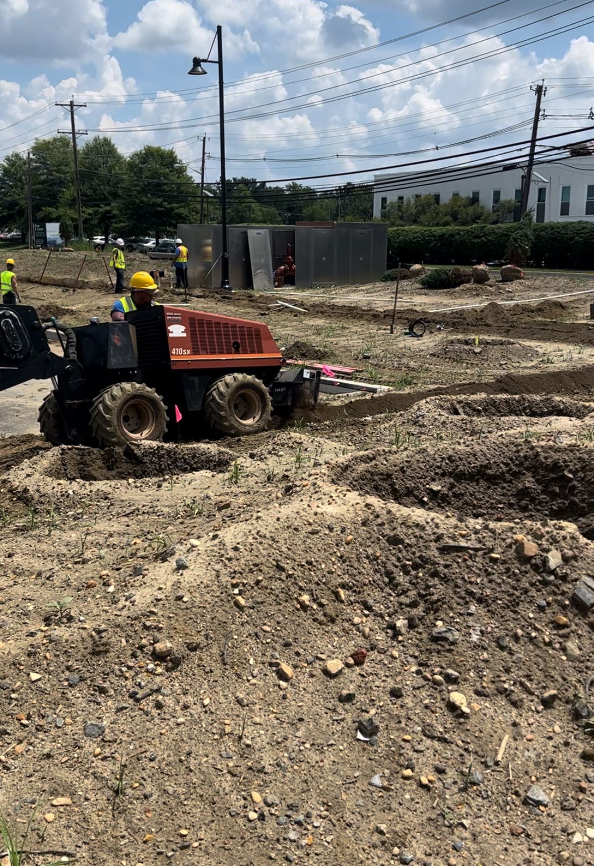 Construction crew operating a small loader on a muddy, gravel site under a cloudy sky