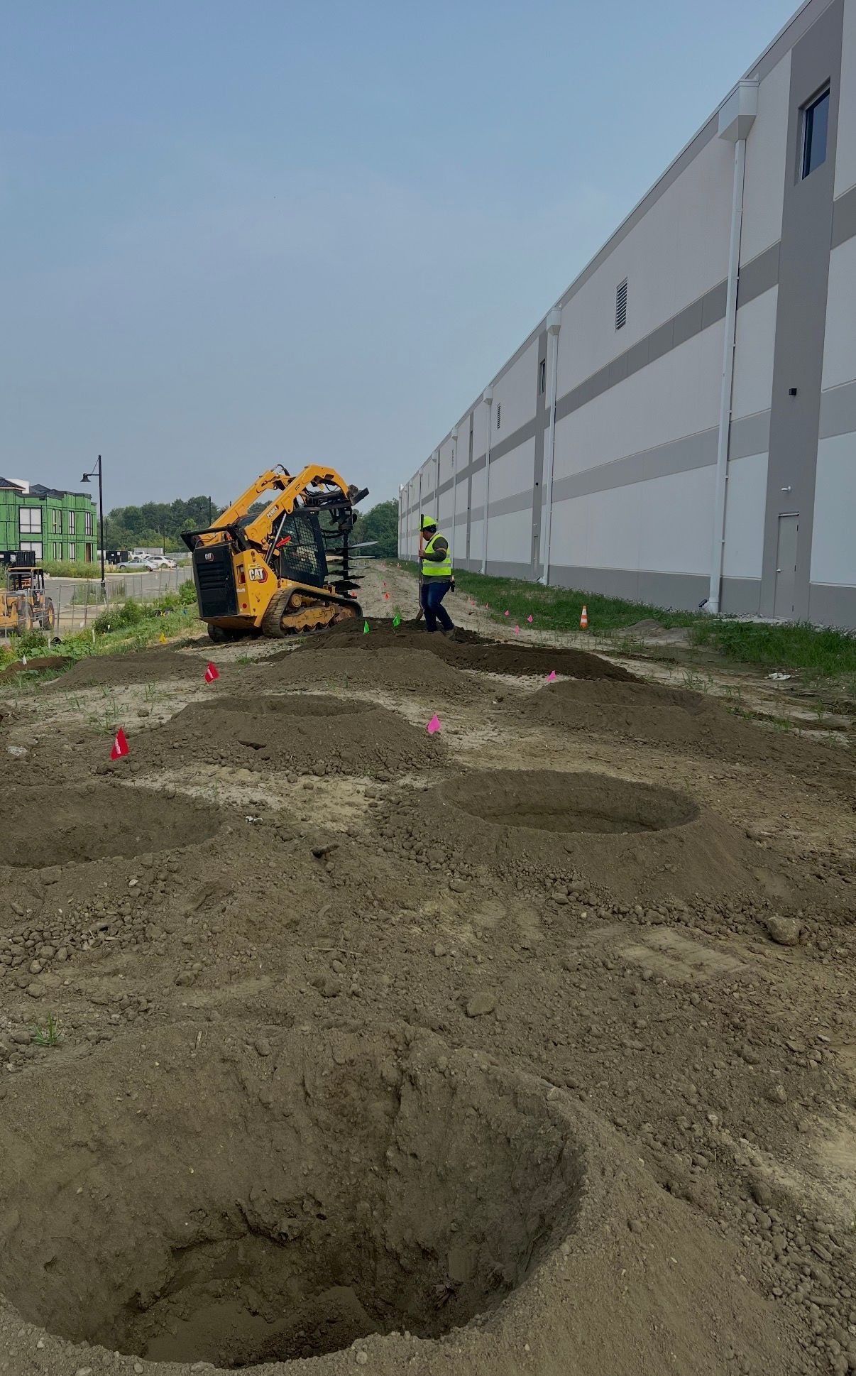 Construction worker inspecting a trench beside a long concrete wall, with a yellow machine nearby.