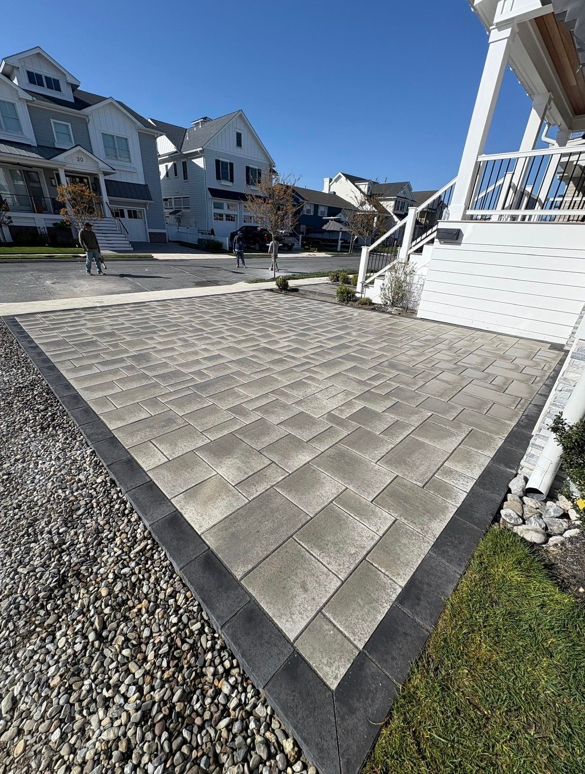 Paver patio beside a white house, bordered by dark edging and gravel in a sunny coastal neighborhood.