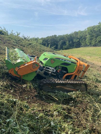 Green agricultural machine tipped on its side in a muddy field beside a grassy embankment