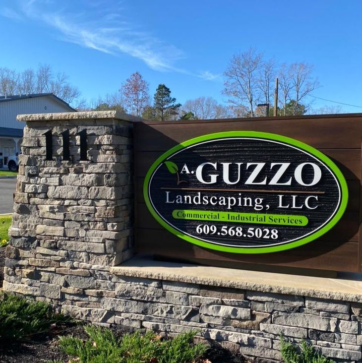 A Guzzo Landscaping LLC sign on a stone wall under a blue sky