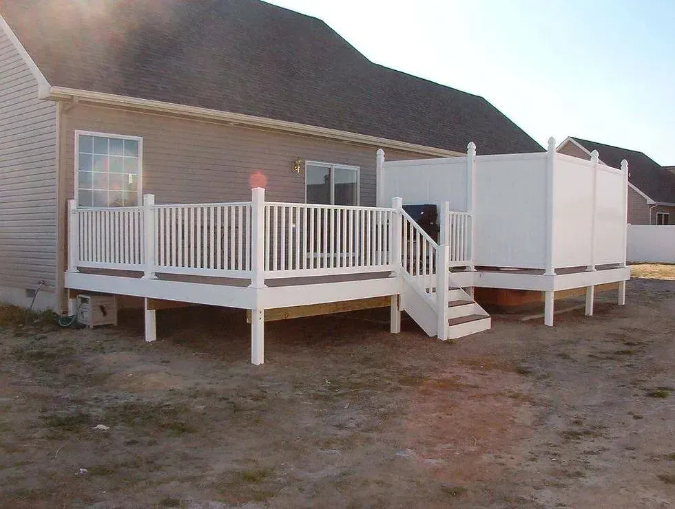 White wooden deck and stairs attached to the back of a beige house in a dirt yard.