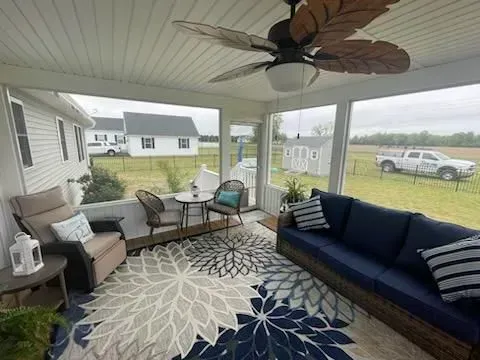 Covered porch with navy sofa, chairs, patterned rug, ceiling fan, and view of a grassy yard with houses.