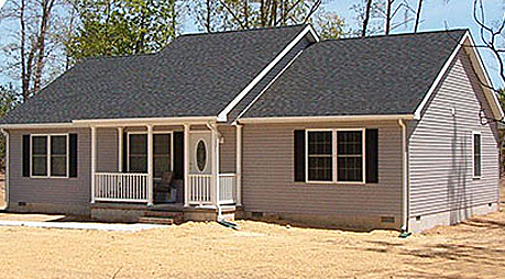 A small house with a porch and a black roof is sitting on top of a dirt field.