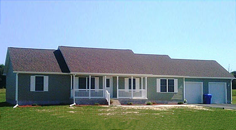 A house with a porch and a garage is sitting in the middle of a grassy field.