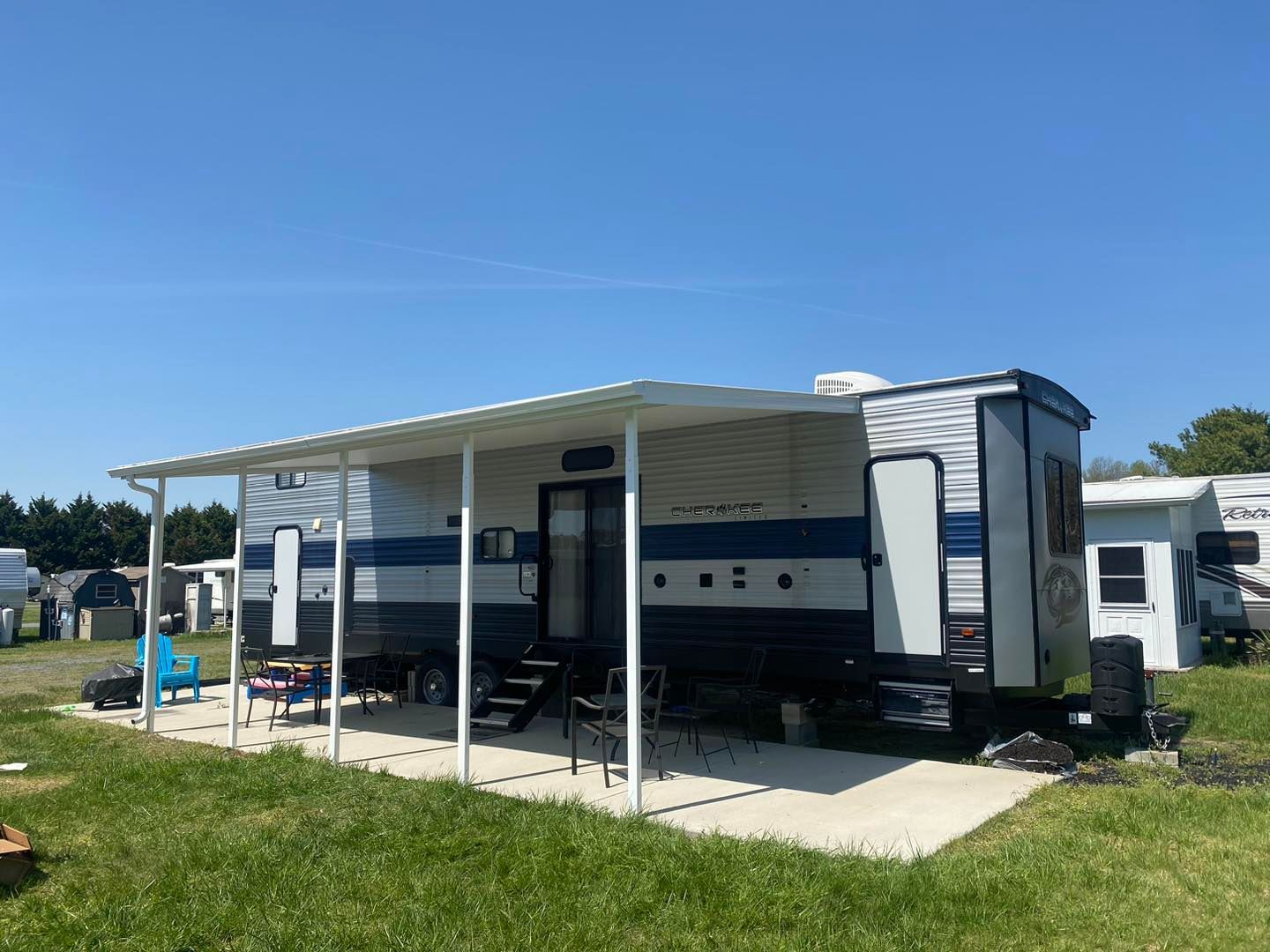 A blue and white trailer with a covered porch is parked in a grassy field.