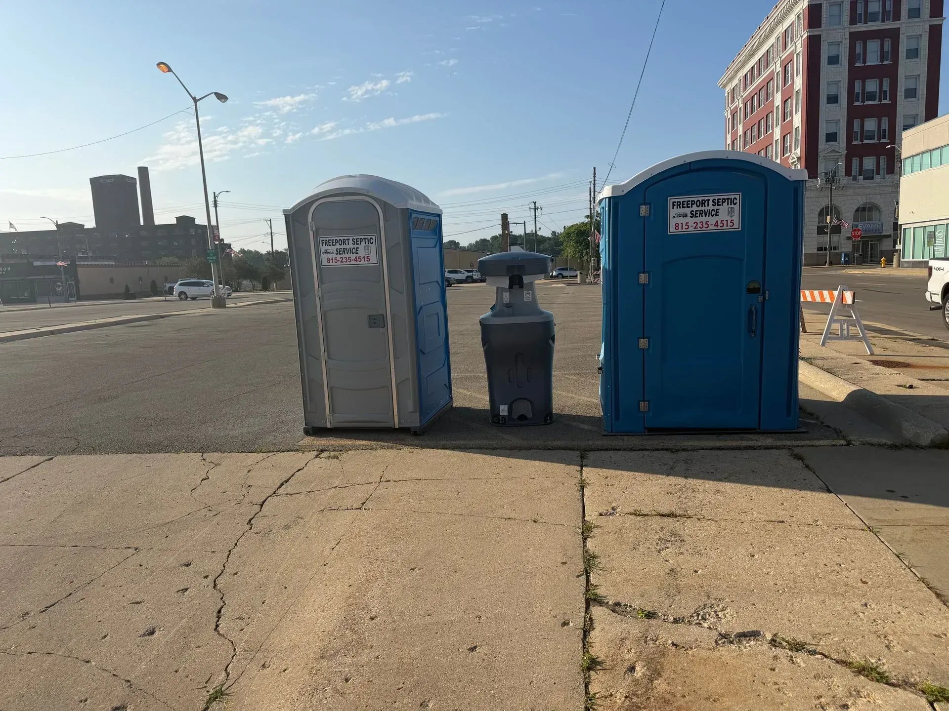 Blue and gray portable toilets beside a trash bin on a sunny city street corner.
