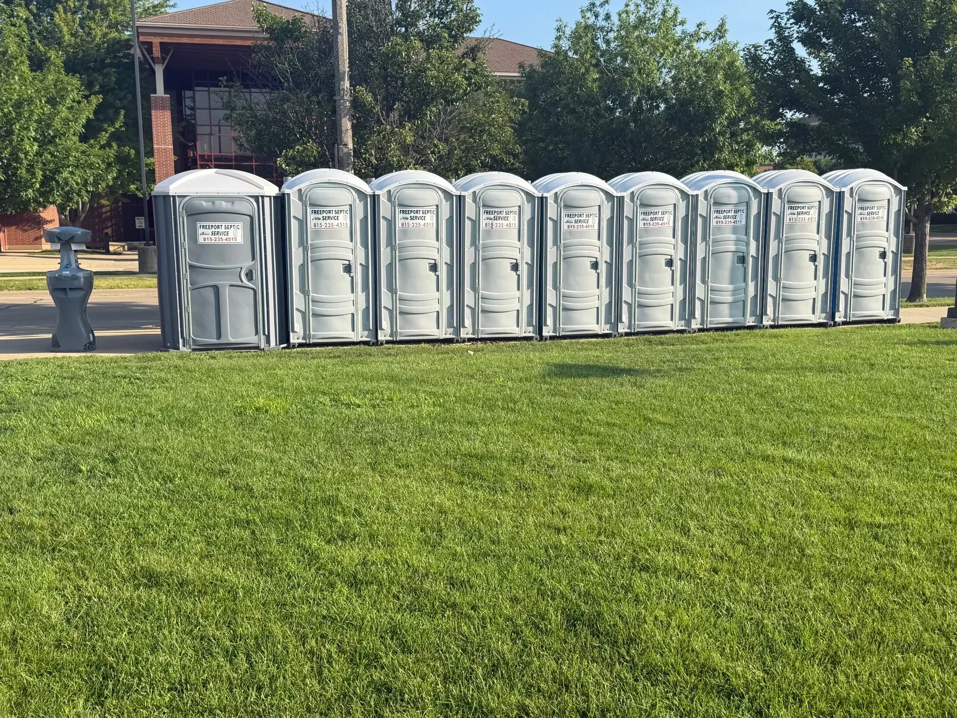 Nine gray portable toilets lined up on a grassy lawn beside a street and houses.