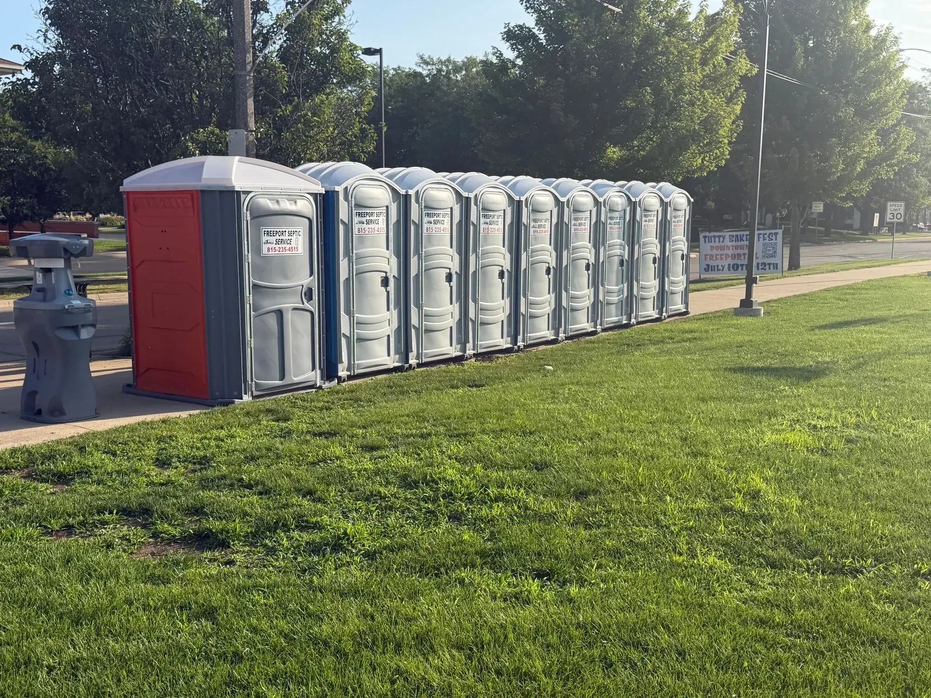 Row of gray portable toilets and a red unit beside a grassy park path on a sunny day.