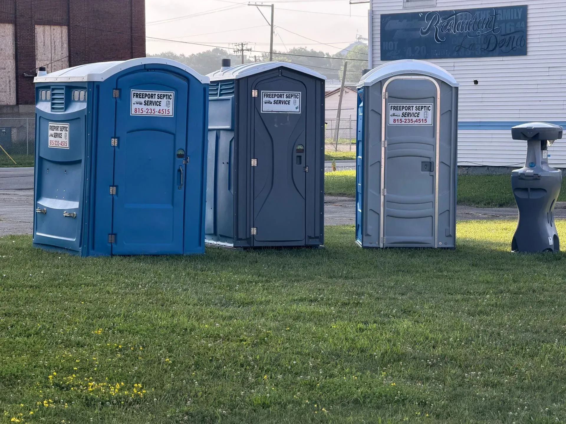 Three portable toilets—two blue and one gray—on grass near a building in winter.