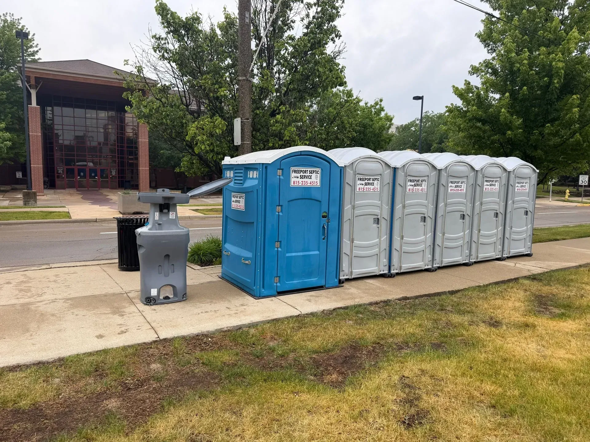 Portable toilets and a trash can beside a sidewalk near a brick building.