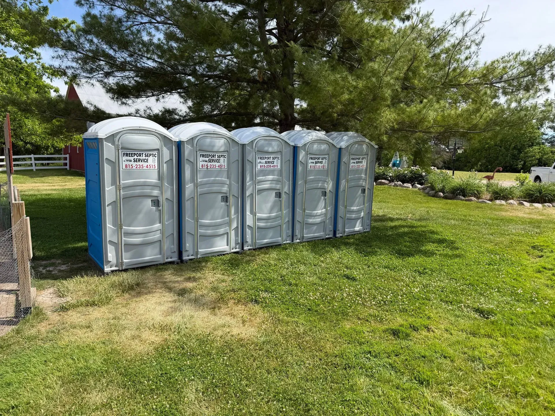 Four portable toilets beside a grassy park area with trees and a sidewalk.