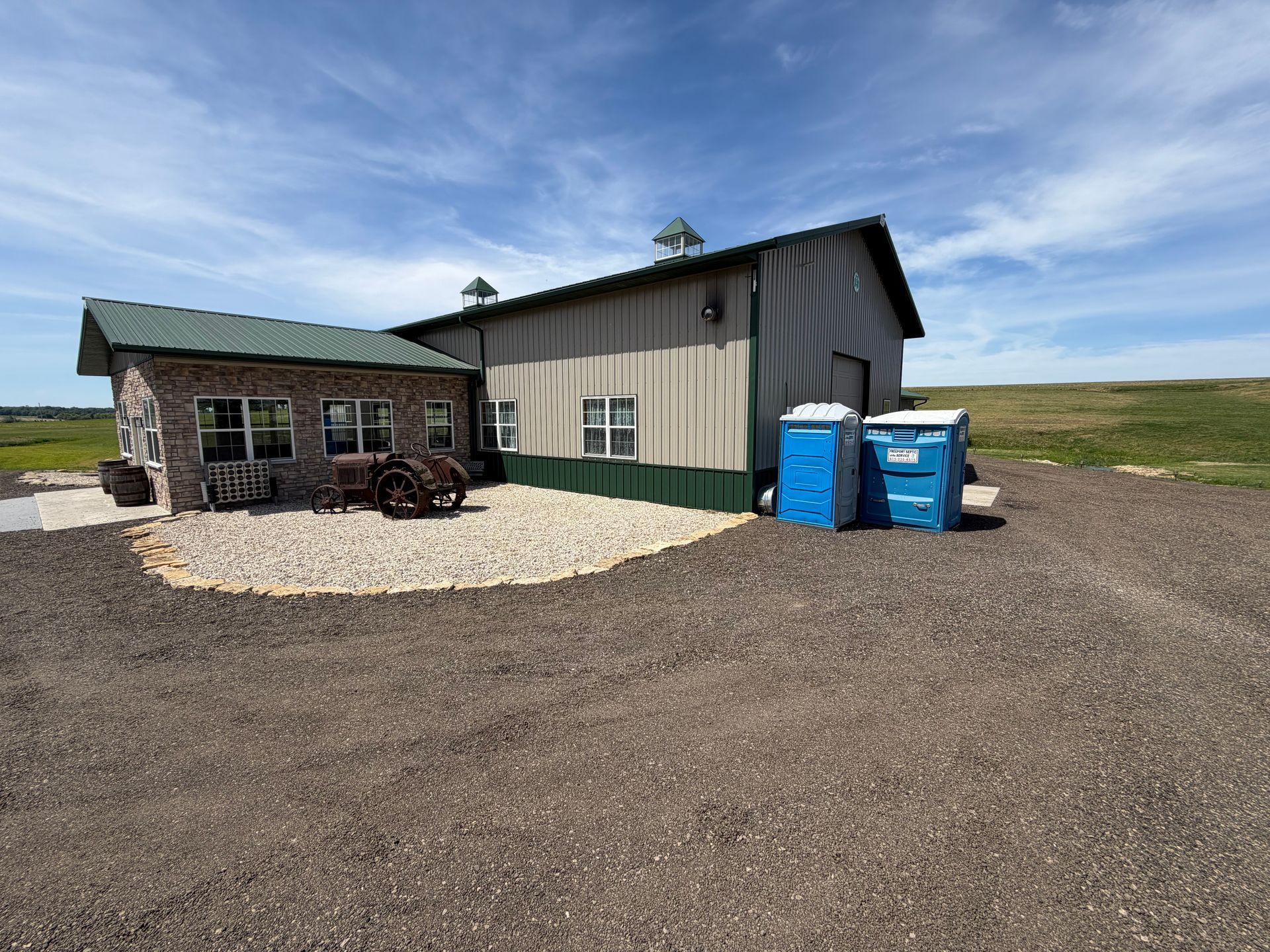 Rural building with stone front and metal shed, blue trash bins, and a wagon on gravel under a cloudy sky