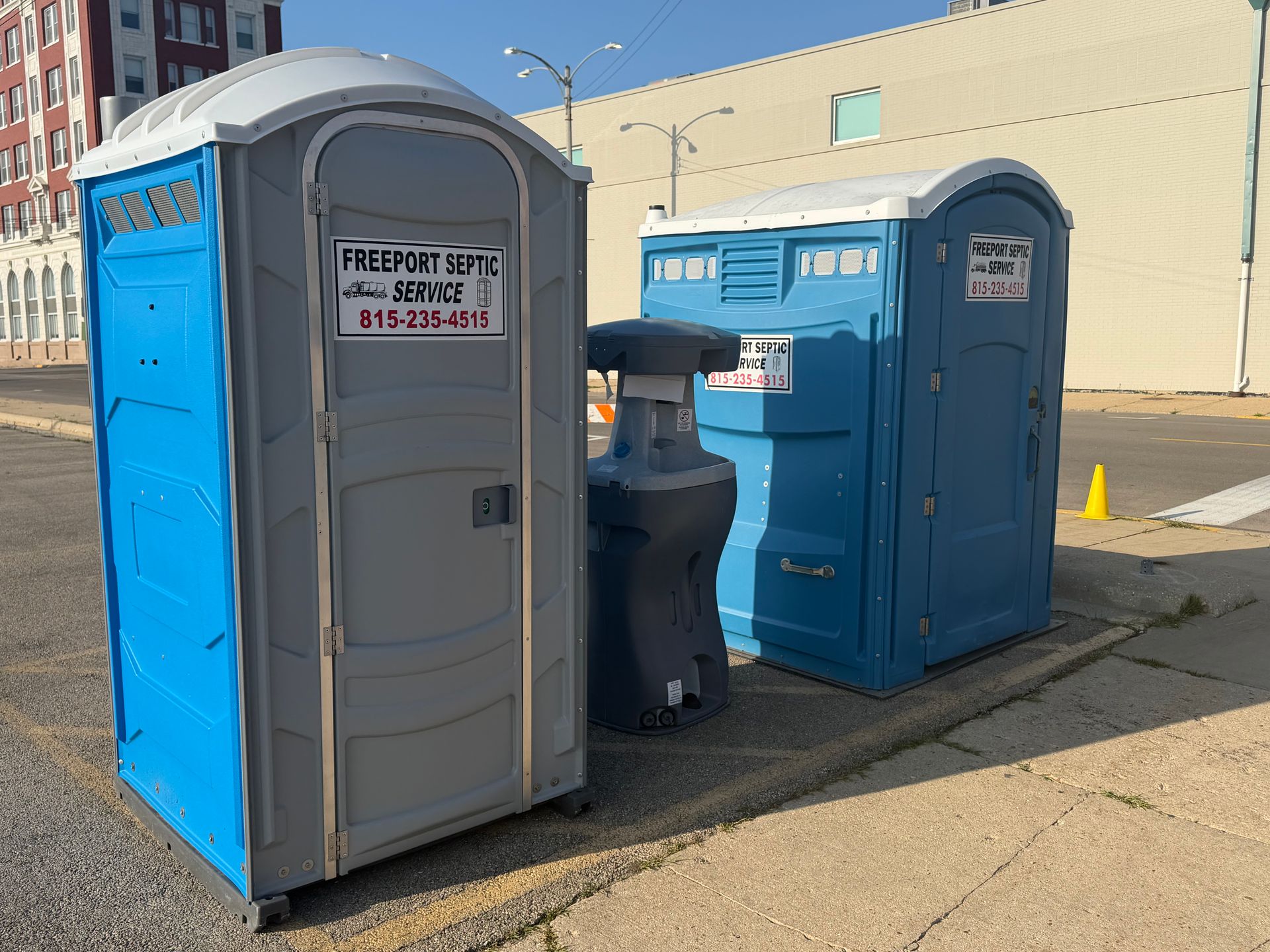 Two blue portable toilets and a gray trash can on a sunny sidewalk beside a building