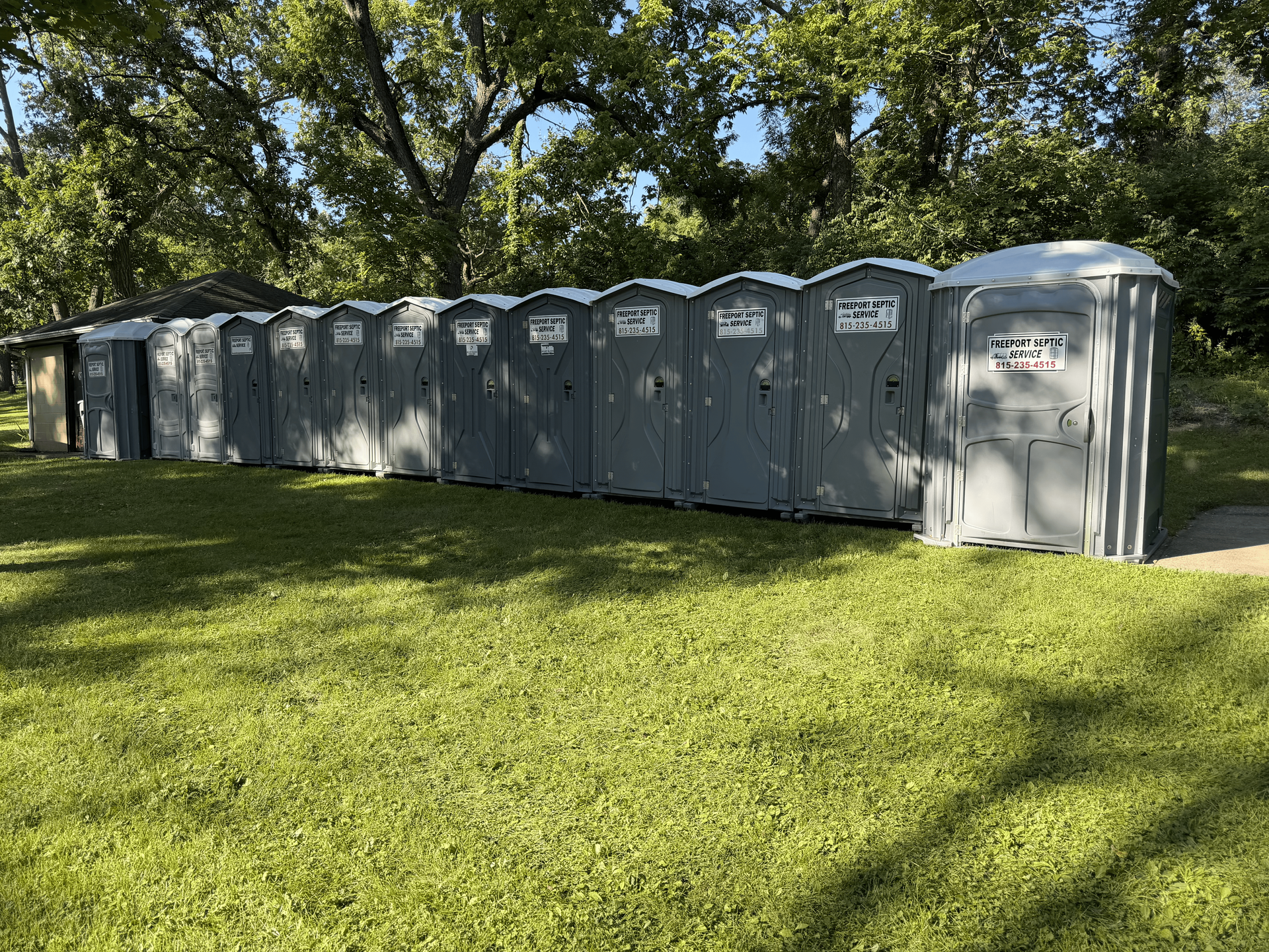 A row of gray portable toilets on a grassy lawn beside trees in sunlight