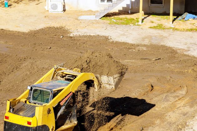 Yellow toy dump truck digging in a dirt pile near a house with a staircase and air conditioner.