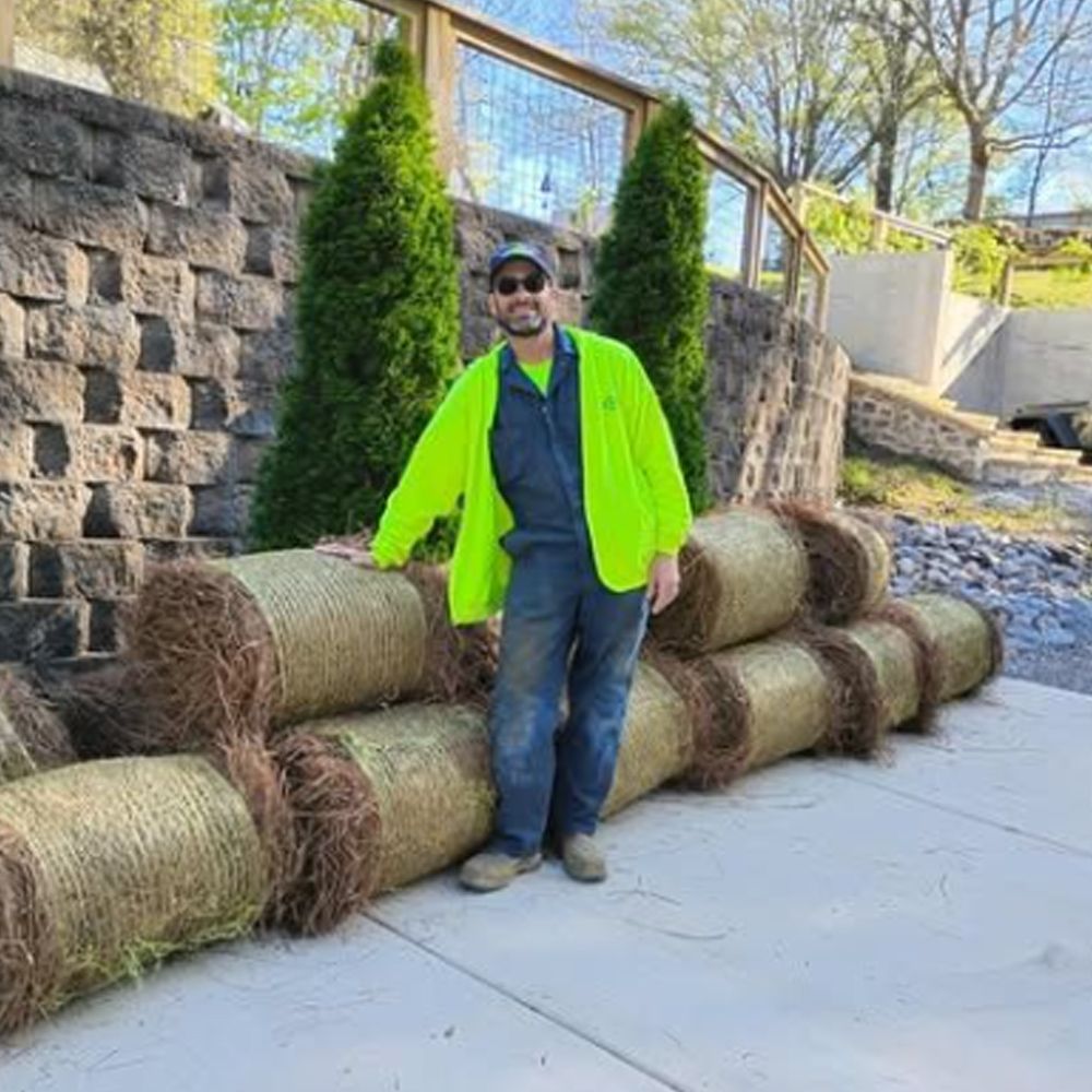A man in a neon yellow jacket is standing next to a pile of pine straw.
