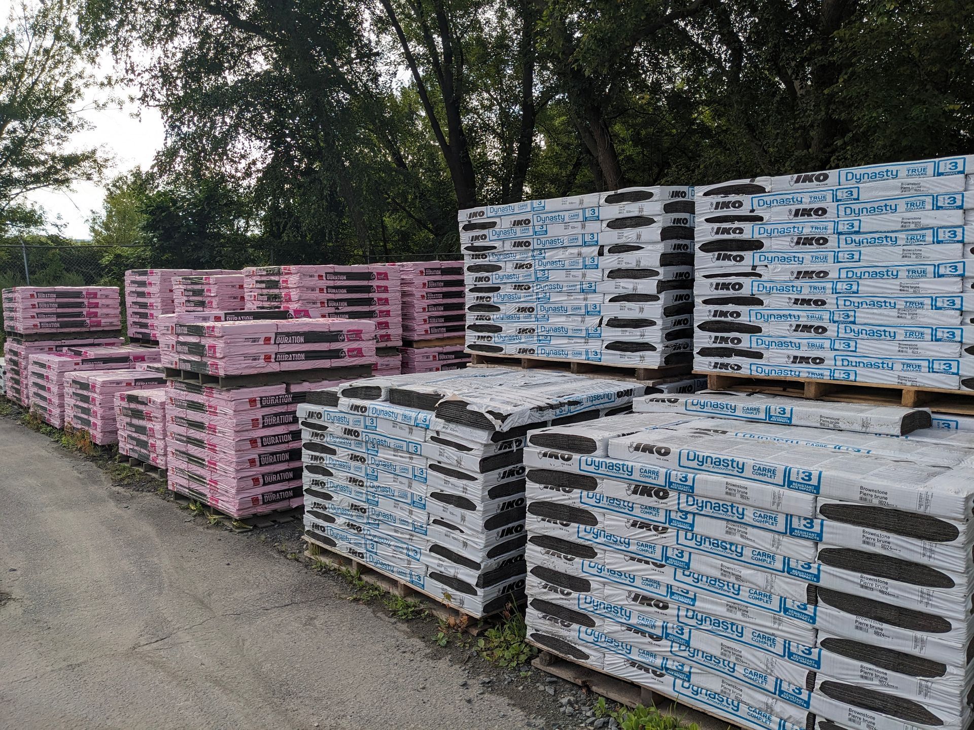 Stacks of roofing shingles on pallets; pink and blue packaging outdoors.