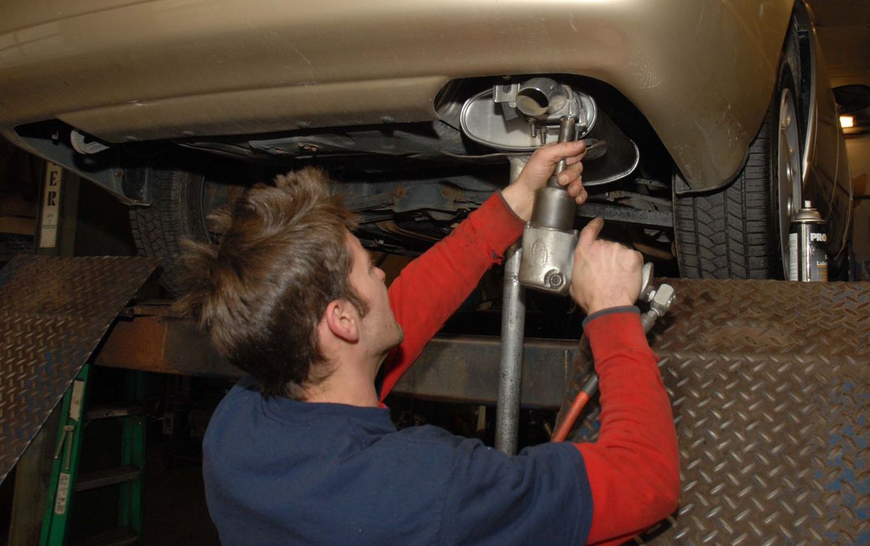 Mechanic working under a car on a lift, using a tool on the exhaust system. Indoors.