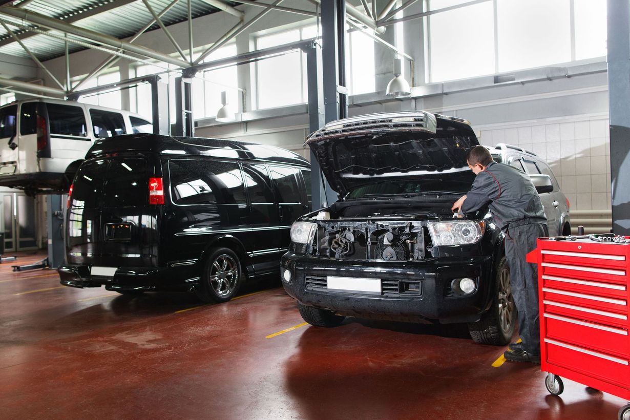 Mechanic working on a black truck in a car repair shop. Other vehicles and a toolbox are nearby.