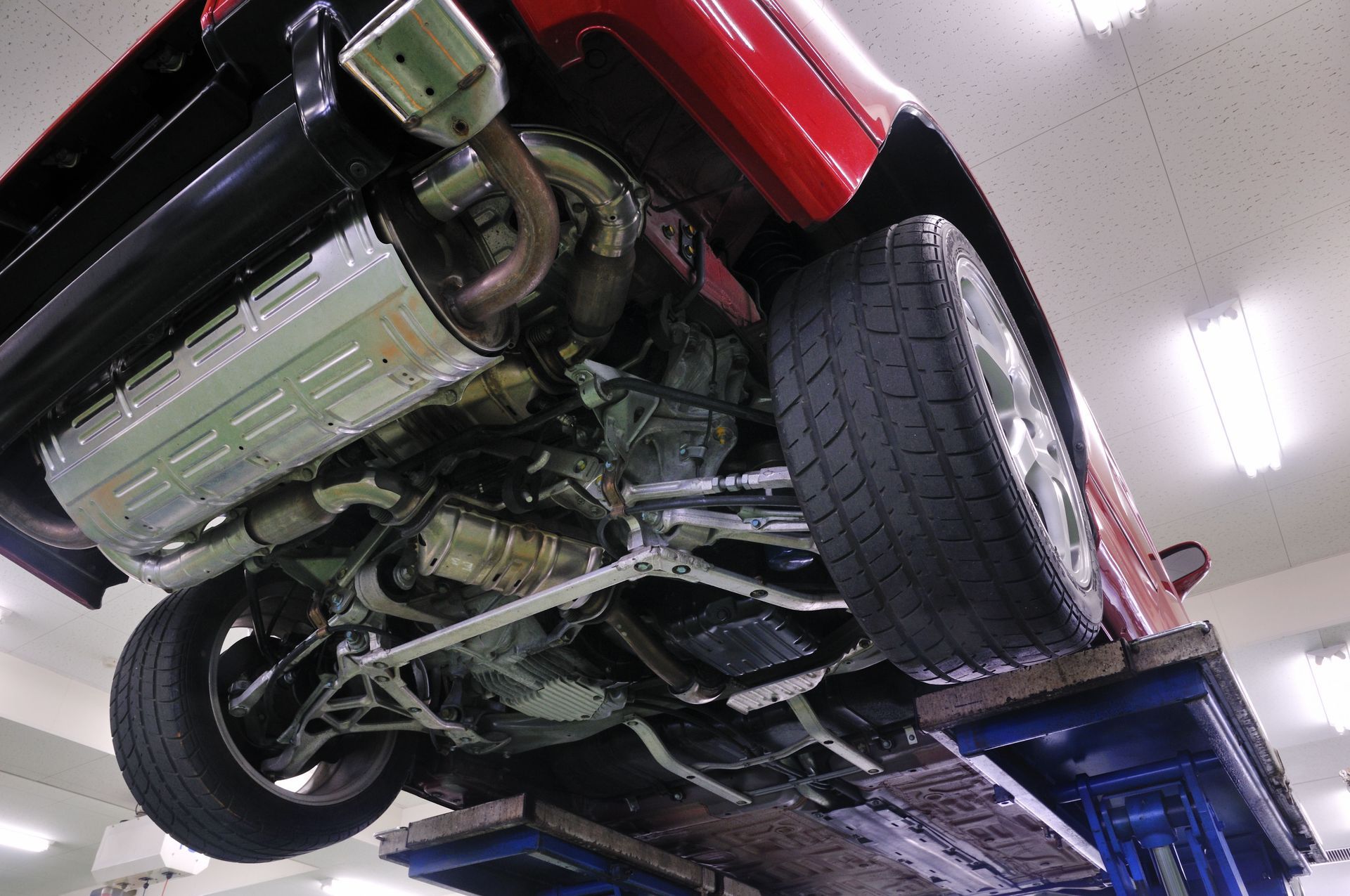 Underside of a red car on a lift in a garage, showing exhaust system, suspension, and tires.
