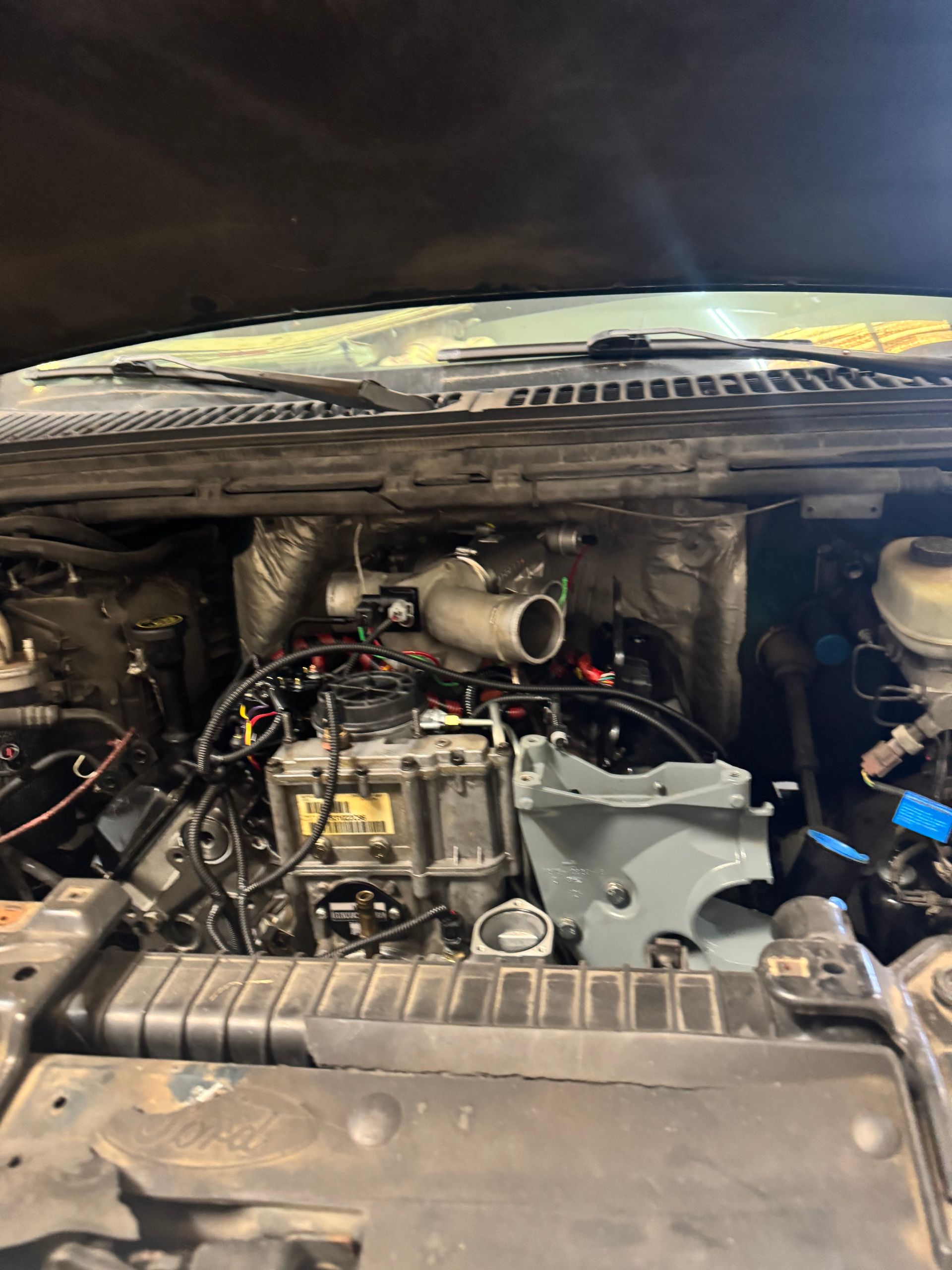 Open car engine bay with exposed parts and hoses, viewed from the front.
