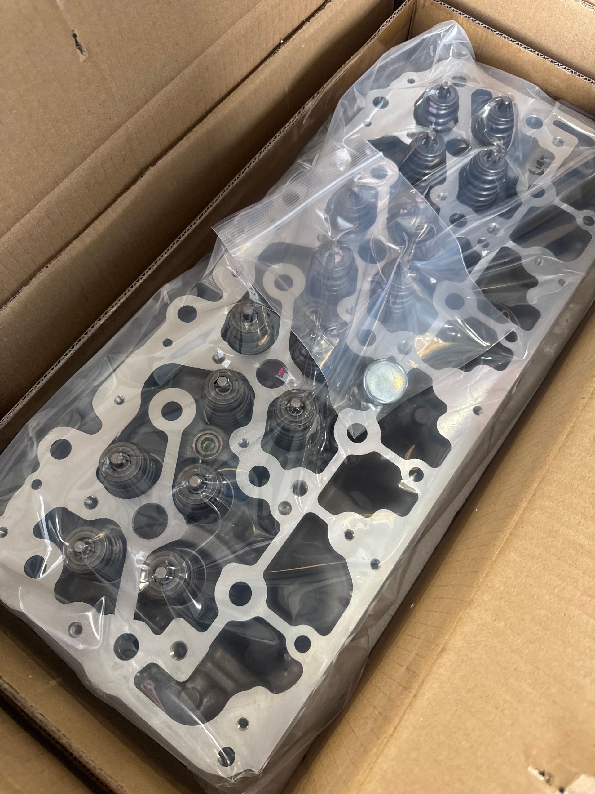 Engine cylinder head in a cardboard box, wrapped in clear plastic.