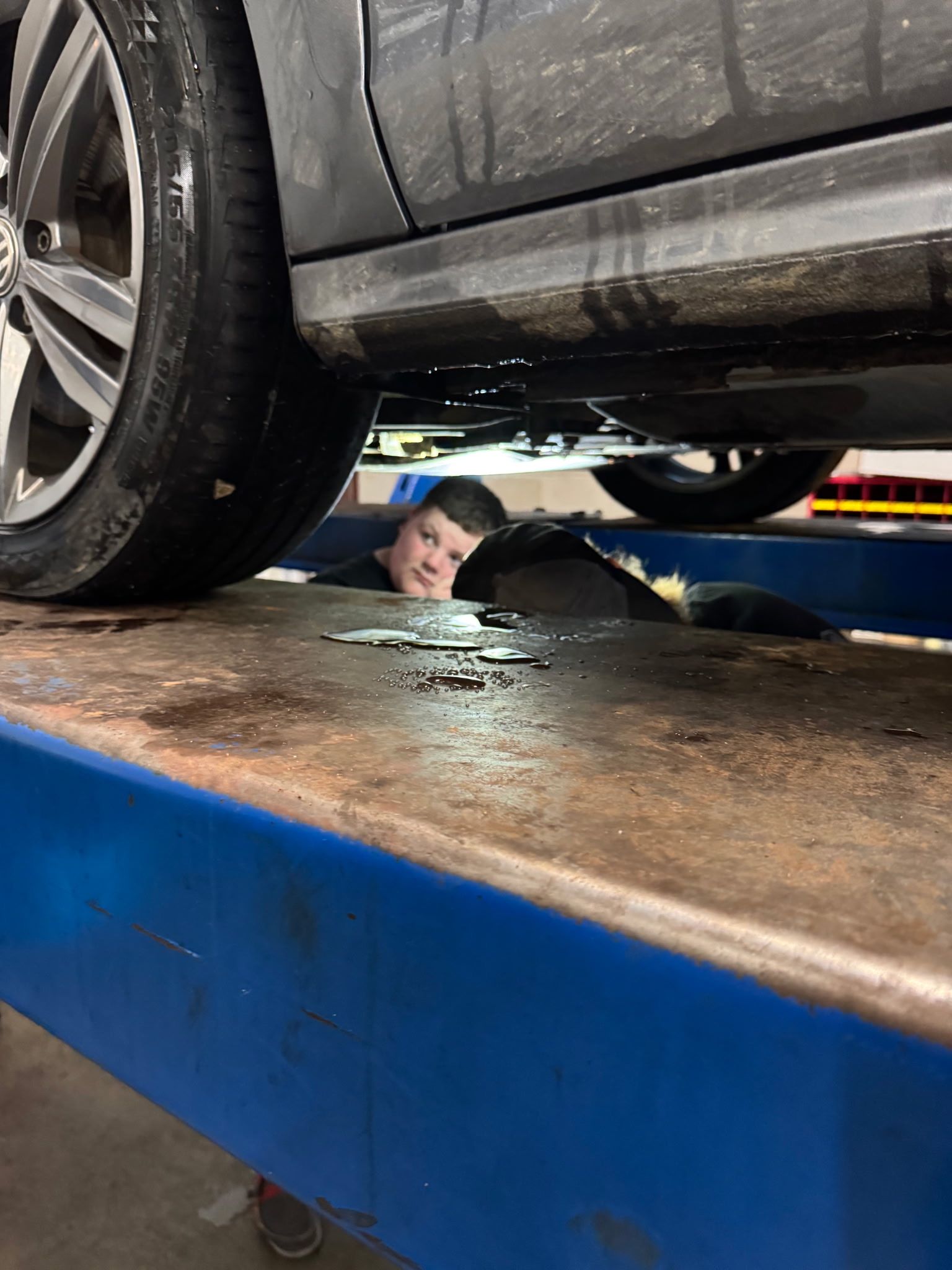 Man under a car on a blue lift in an auto shop, inspecting the underside.