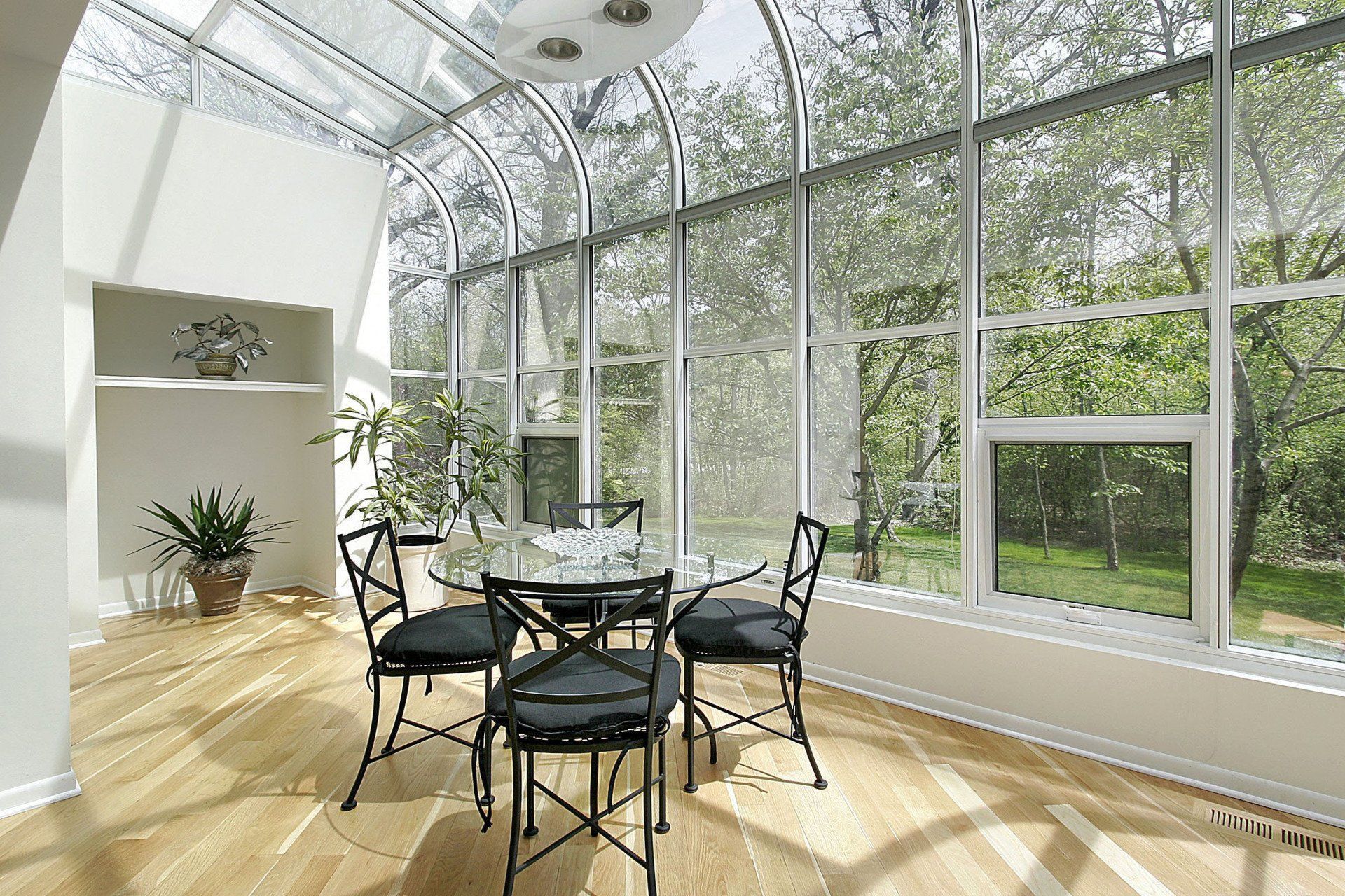 Bright sunroom with a round dining table, black chairs, and large glass windows overlooking greenery