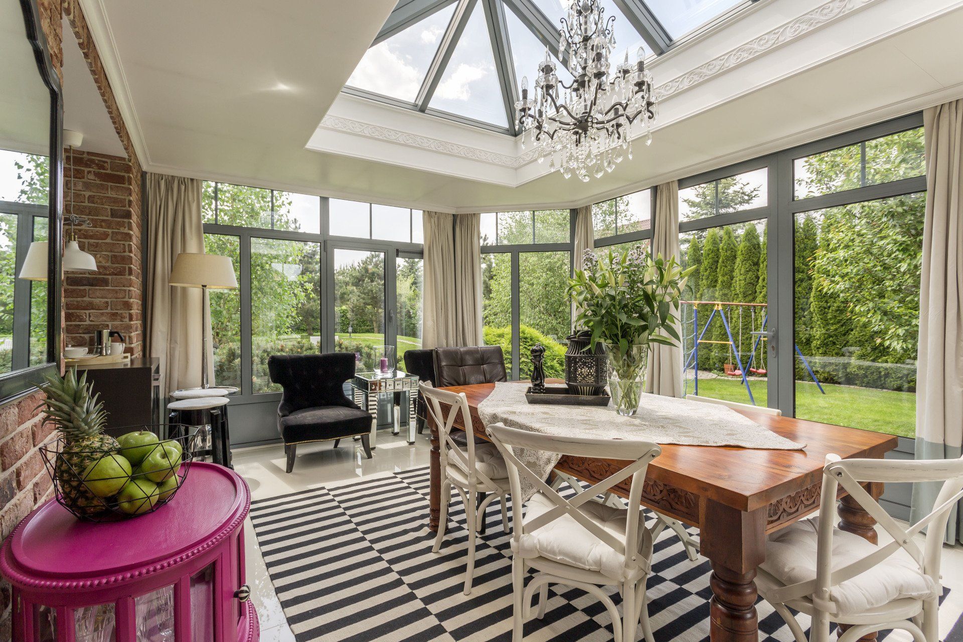 Bright sunroom dining area with large windows, striped rug, white chairs, and plants overlooking a garden