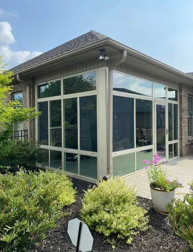 Glass-enclosed sunroom on a house patio, surrounded by shrubs and potted flowers under a blue sky