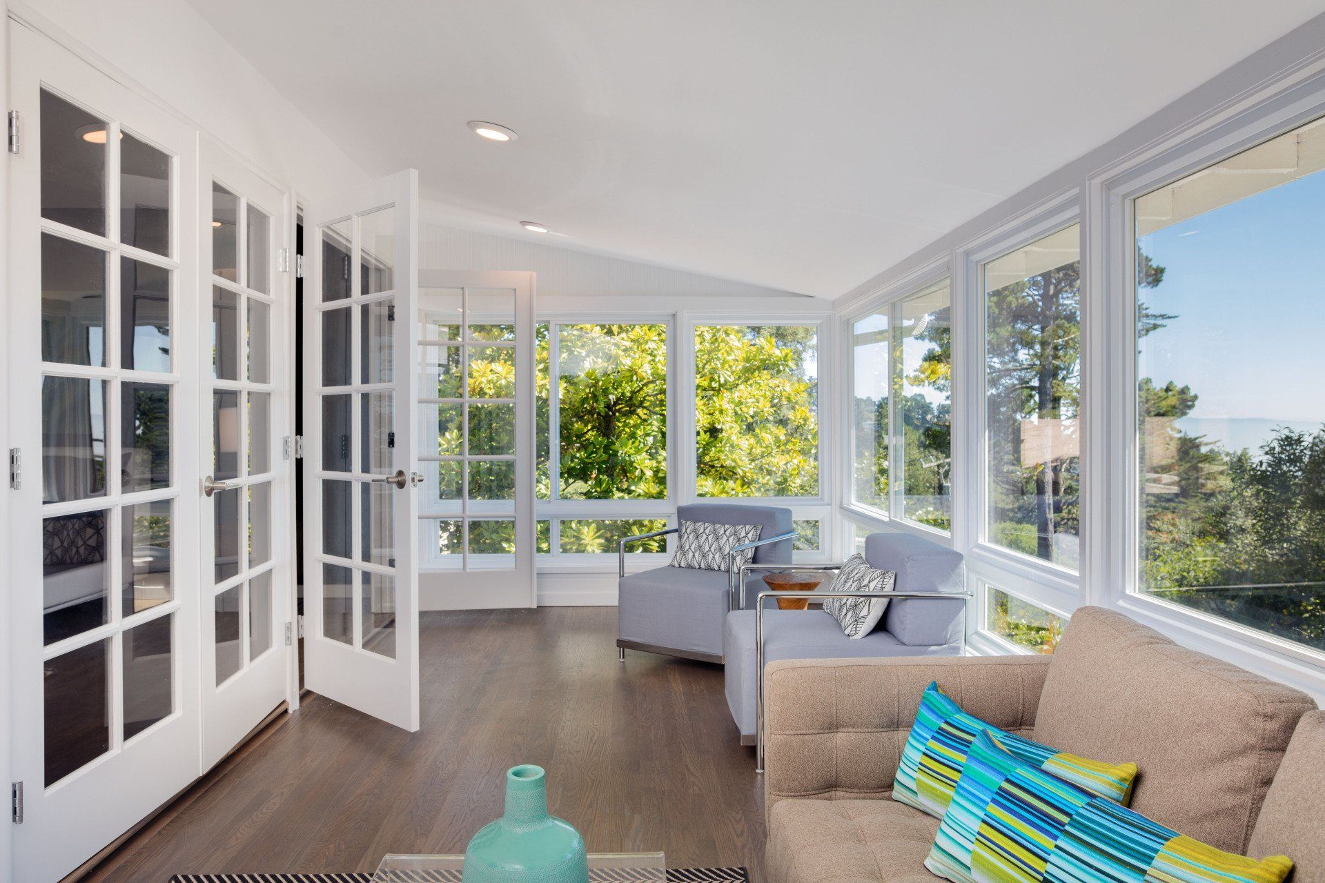 Bright sunroom with white French doors, wicker seating, and large windows overlooking greenery